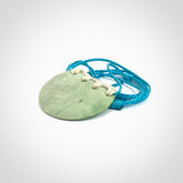 Hand carved Waimea pounamu disc pendant. These beautiful pieces have been carved for us by Raegan Bregmen and are quite beautiful. The soft green colours are lovely and are set off with a paradise blue cord and ice white ridge binding.
